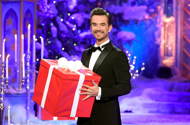 Florian Silbereisen lädt ein zum „Adventsfest der 100.000 Lichter“. Florian Silbereisen lacht und hält ein großes rotes Paket in den Händen