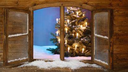 Ein Fenster steht offen und gibt den Blick auf einen beleuchteten Weihnachtsbaum und eine Schneelandschaft frei.